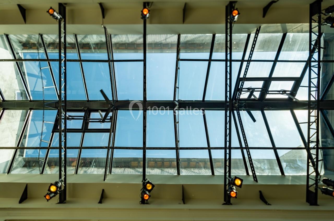 Vue en contre-plongée d'une verrière moderne avec des structures métalliques et des projecteurs fixés.