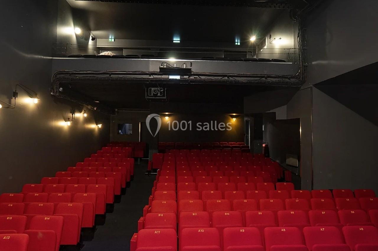 Salle de théâtre avec rangées de sièges rouges vides et un balcon supérieur dans un éclairage tamisé.