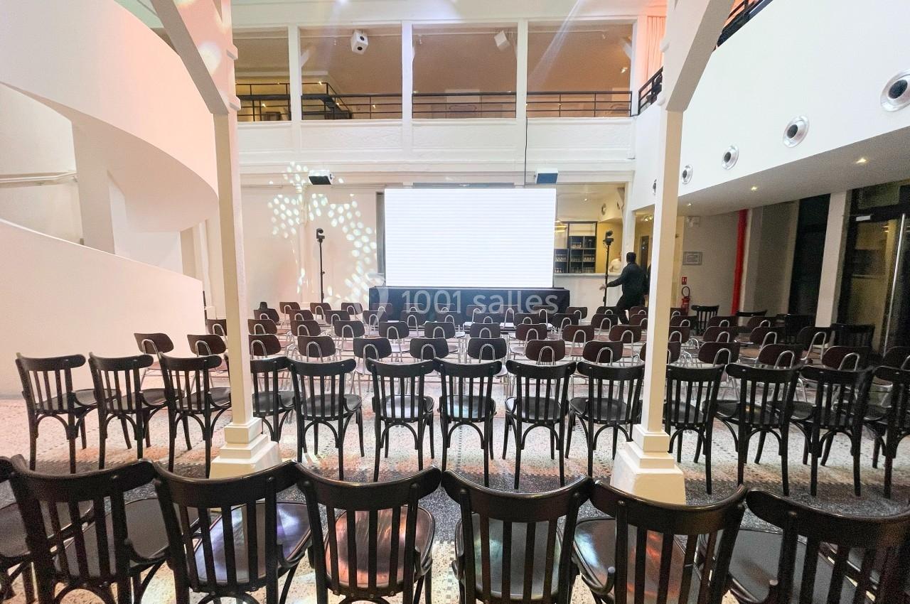 Salle de conférence avec des rangées de chaises noires face à un écran blanc, éclairage tamisé.