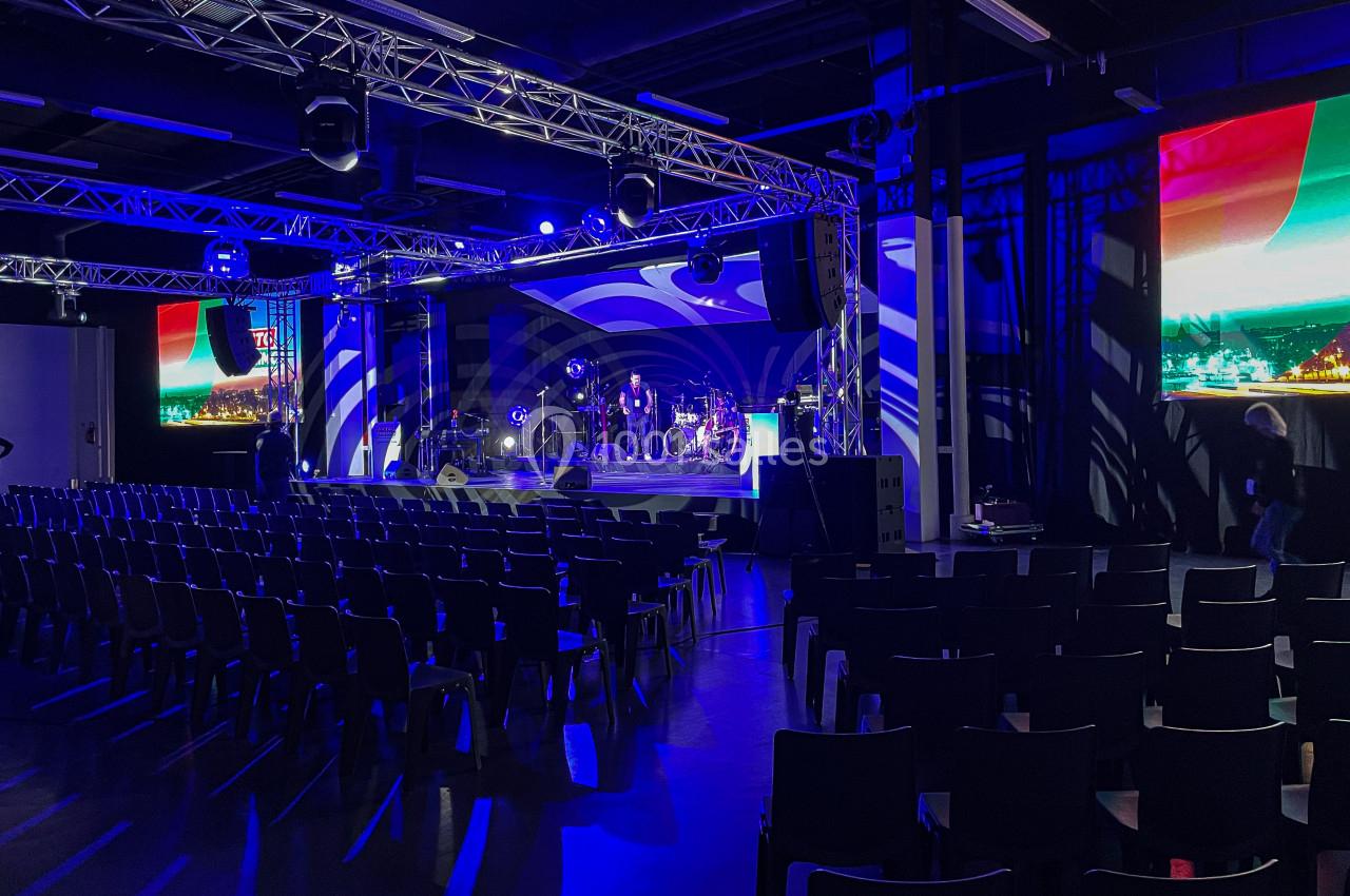 Salle de spectacle avec scène éclairée en bleu, chaises noires alignées et écrans affichant des images colorées.