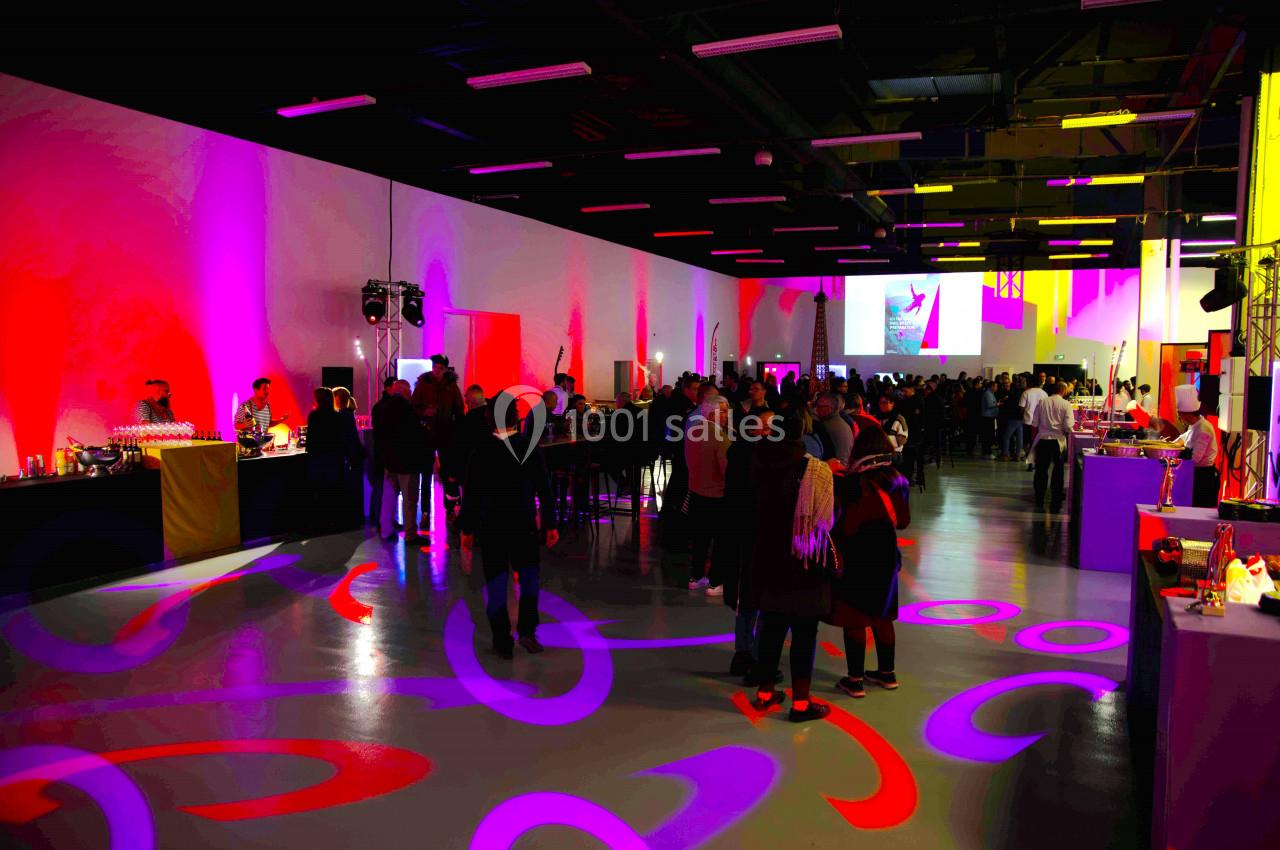 Salle événementielle avec éclairage coloré, tables hautes, buffet et invités discutant dans une ambiance festive.