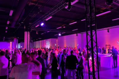 Des personnes discutent et prennent un verre autour de tables en bois devant un bâtiment industriel lors d'un événement.