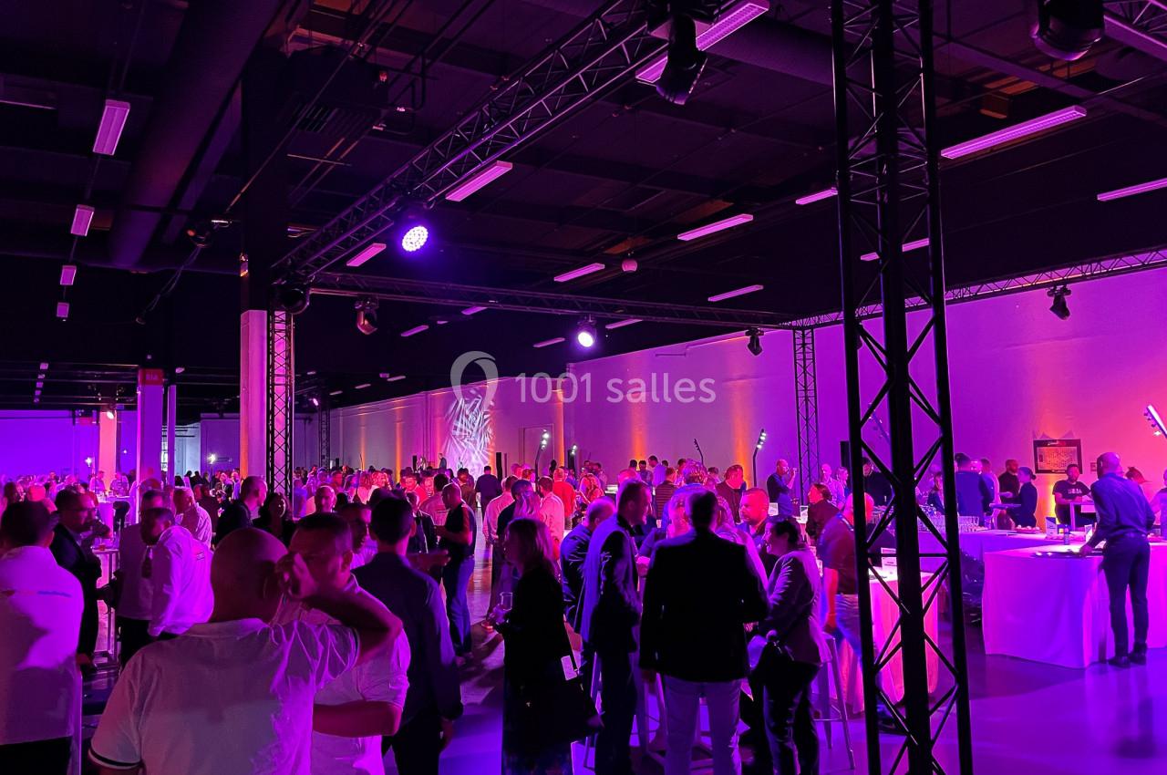 Salle de réception bondée avec des invités debout sous un éclairage violet, discutant autour de tables hautes.