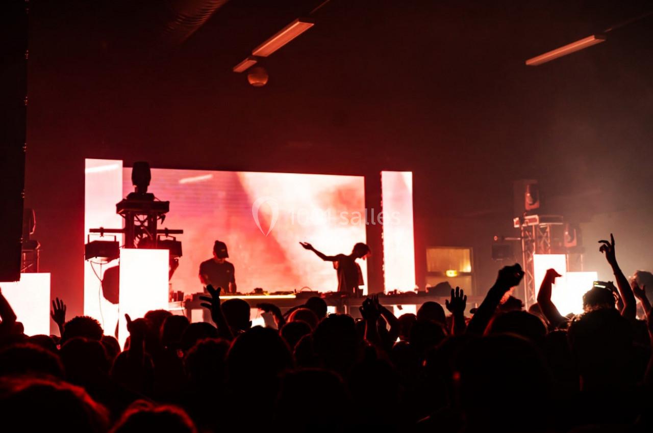 Foule dans une salle de concert sombre, éclairée par des écrans lumineux et un DJ sur scène.