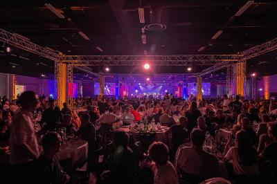 Des personnes discutent et prennent un verre autour de tables en bois devant un bâtiment industriel lors d'un événement.
