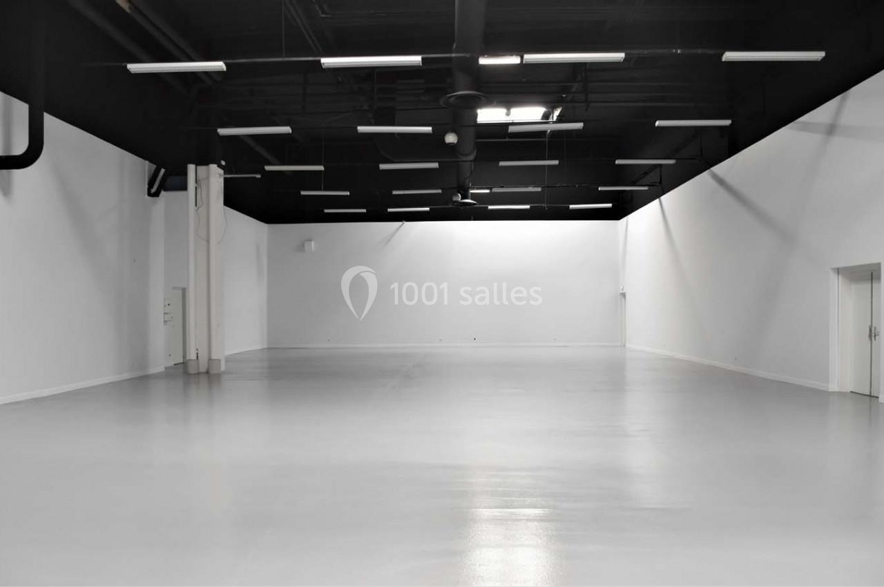 Grand espace intérieur vide avec murs blancs, sol brillant et plafond noir équipé de luminaires suspendus.