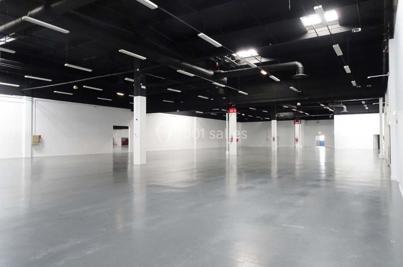 Salle vide avec sol en béton gris, murs blancs et plafond noir équipé de luminaires suspendus.