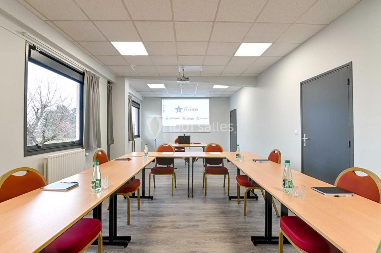 Salle de réunion lumineuse avec tables disposées en U, chaises rouges, bouteilles d'eau et écran de projection.