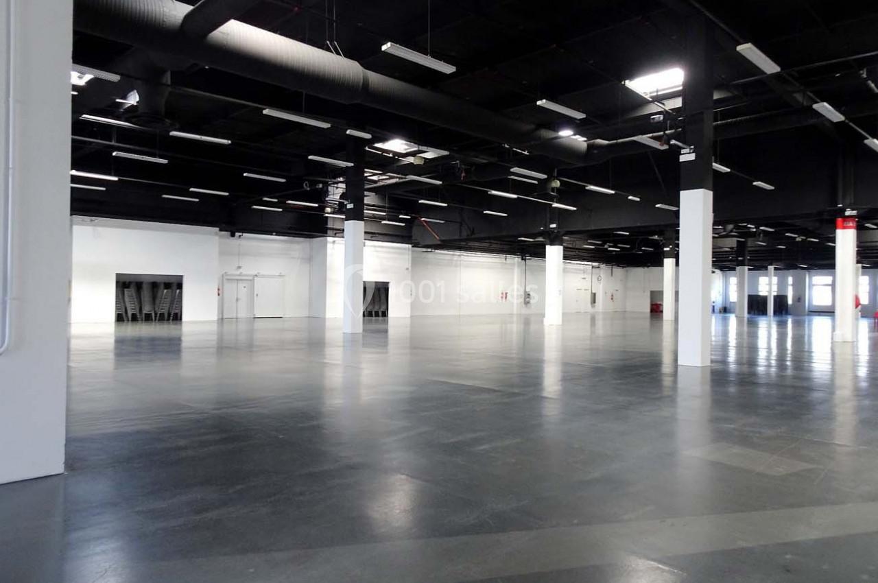 Grand espace intérieur vide avec sol en béton, murs blancs et plafond noir équipé de luminaires suspendus.