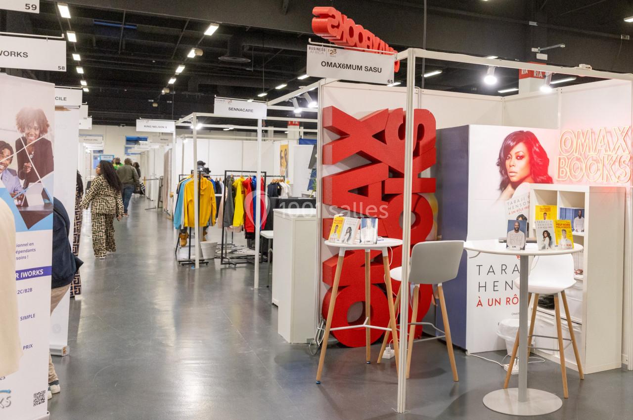 Allée d'un salon professionnel avec stands exposant des livres, vêtements et autres produits sous éclairage intérieur.