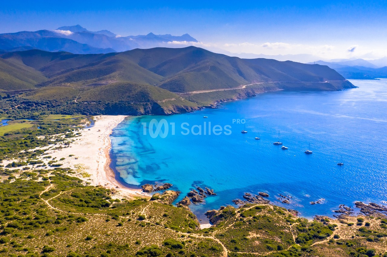 Vue aérienne d'une plage isolée bordée par une mer turquoise, entourée de collines verdoyantes et de montagnes.