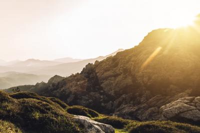 Paysage montagneux au coucher du soleil avec une personne debout sur un promontoire rocheux, entourée de végétation.
