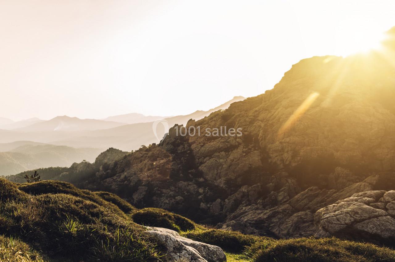 Paysage montagneux au lever du soleil, avec des collines verdoyantes et des rayons lumineux traversant le ciel clair.