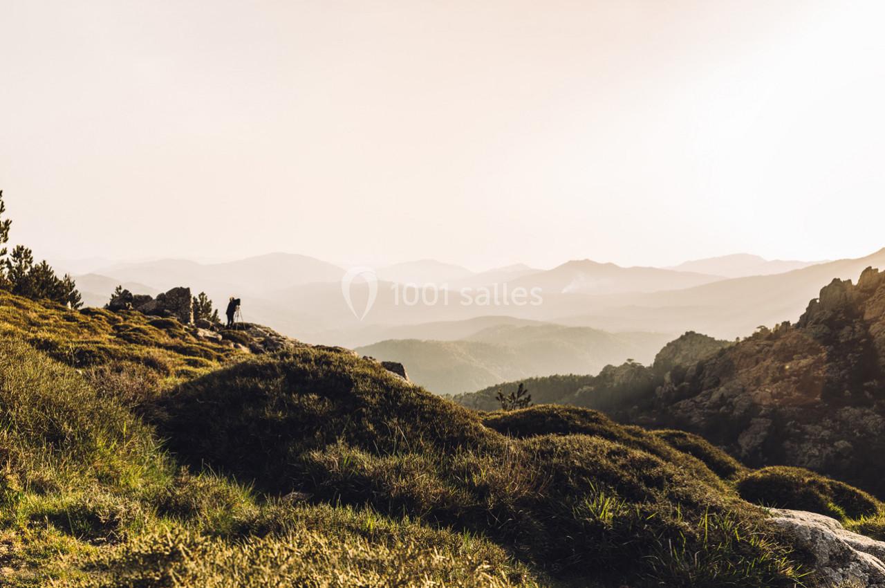 Paysage montagneux au coucher du soleil avec une personne debout sur un promontoire rocheux, entourée de végétation.