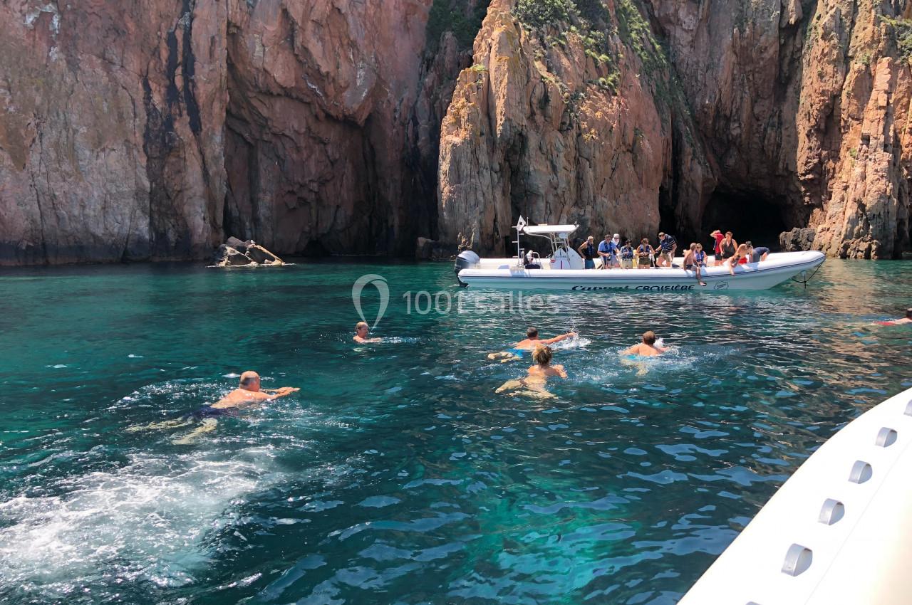Des nageurs dans une eau turquoise près de falaises rocheuses, avec un bateau pneumatique rempli de personnes à proximité.