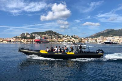 Un bateau pneumatique noir et jaune navigue sur une mer calme, avec une ville et des collines en arrière-plan.