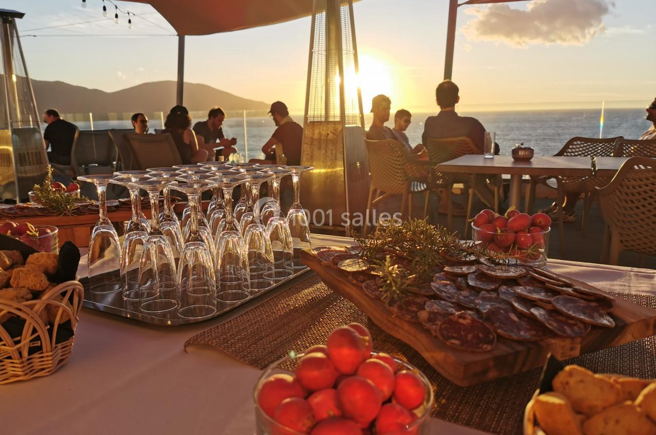 Table dressée avec des amuse-bouches, verres et tomates cerises, en terrasse face à un coucher de soleil sur la mer.