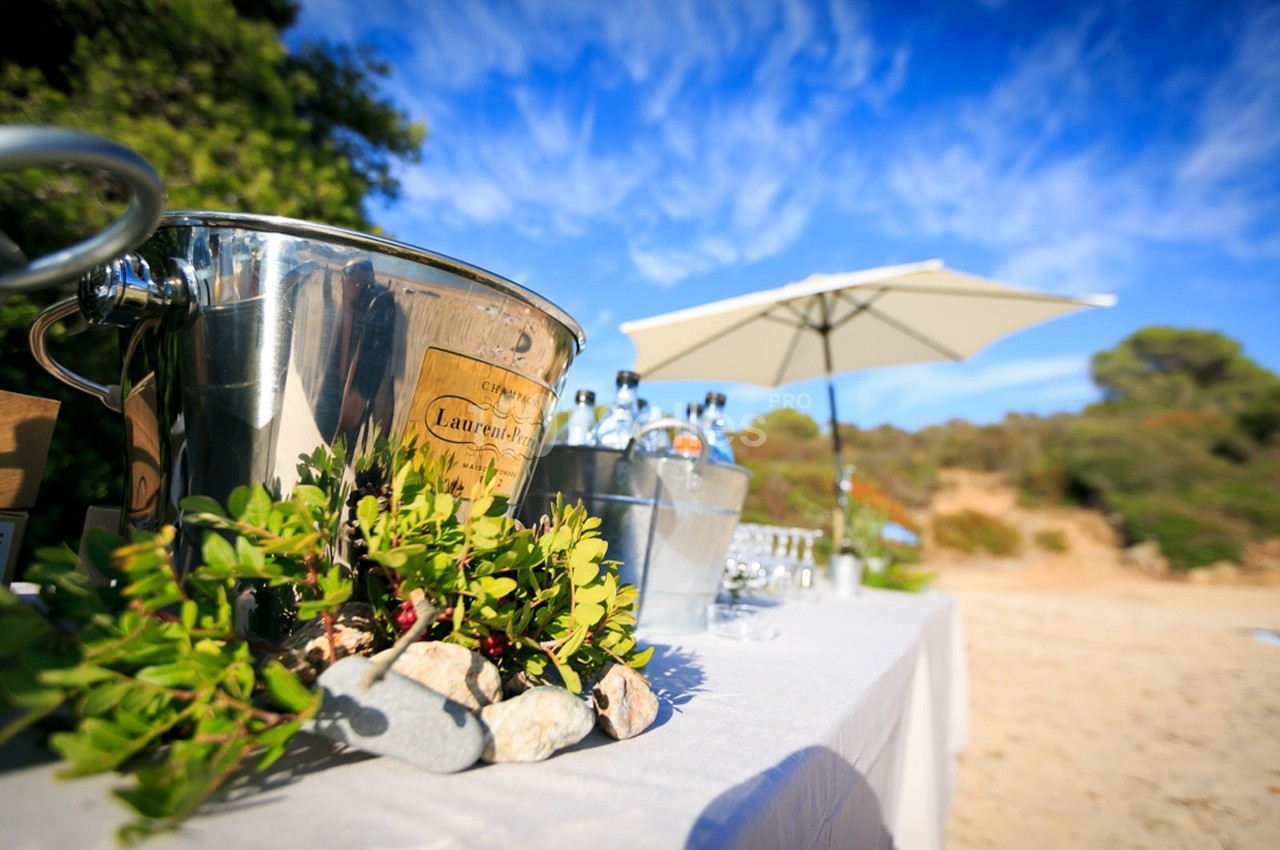 Seau à champagne en métal sur une table blanche en extérieur, avec parasol et végétation en arrière-plan.
