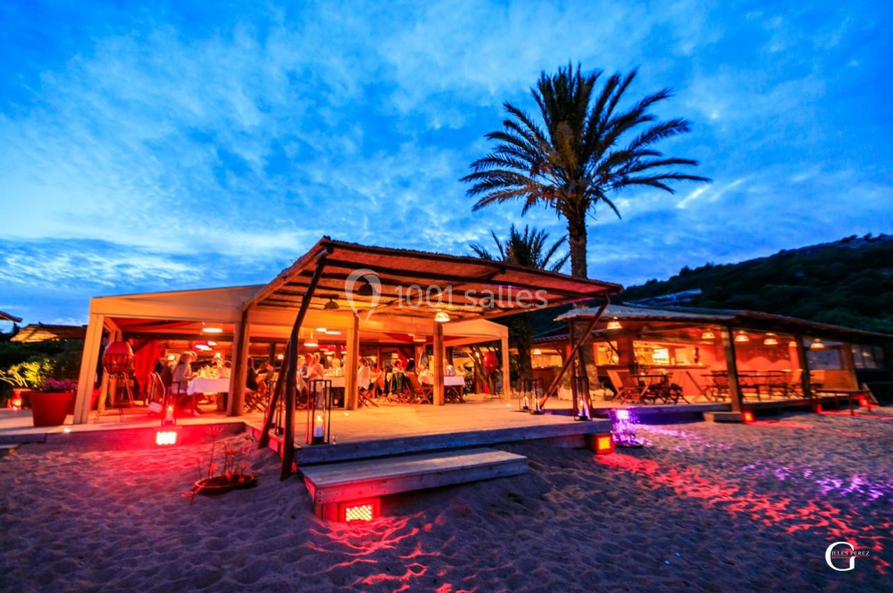 Restaurant en bord de plage avec terrasse éclairée, palmiers et ciel crépusculaire en arrière-plan.