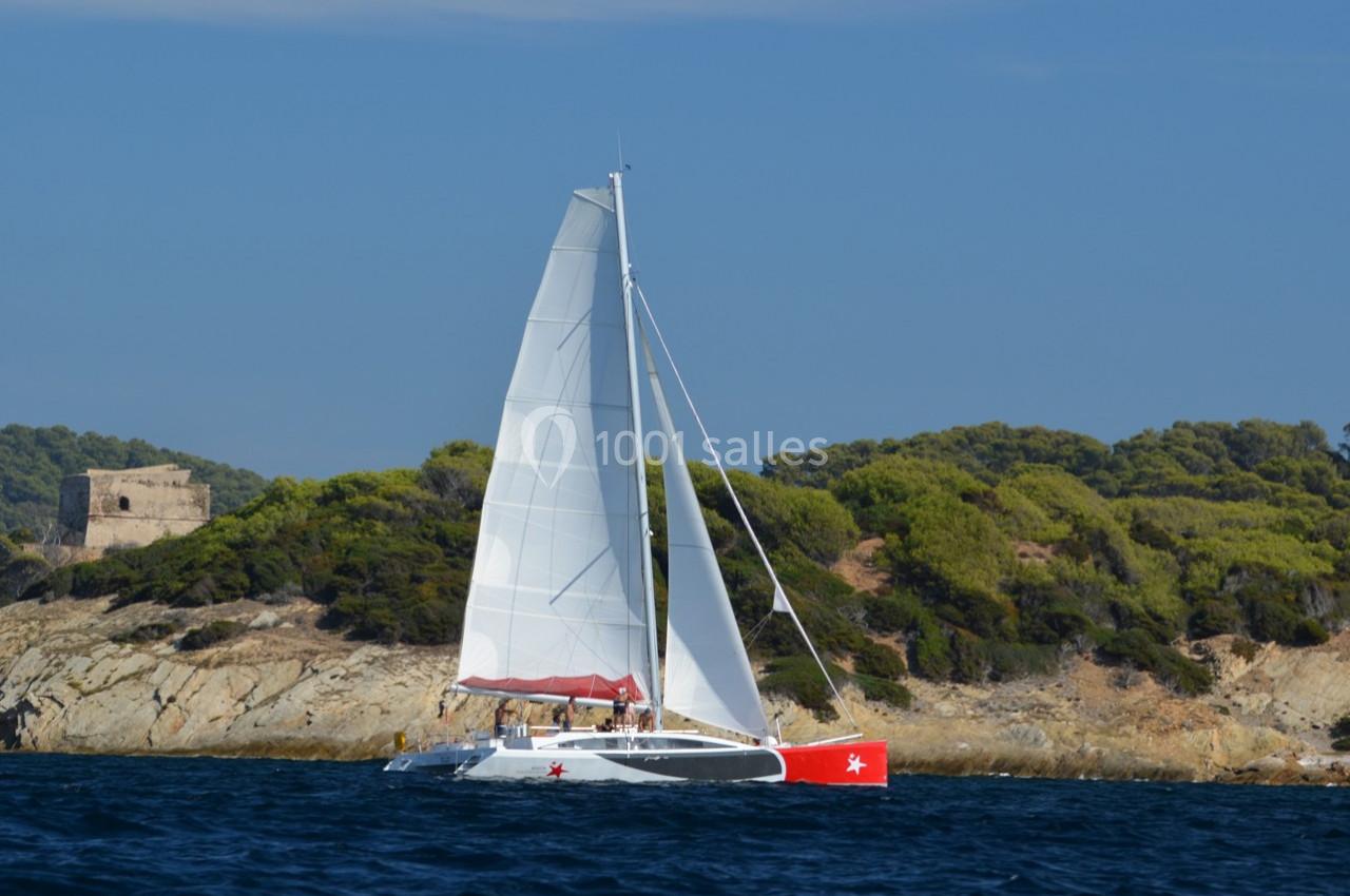 Voilier blanc avec une coque rouge naviguant près d'une côte boisée sous un ciel dégagé.