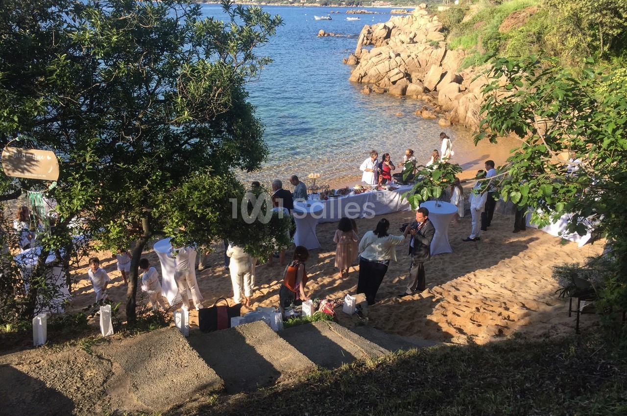 Groupe de personnes rassemblées sur une plage autour de tables dressées, avec vue sur la mer et des collines verdoyantes.