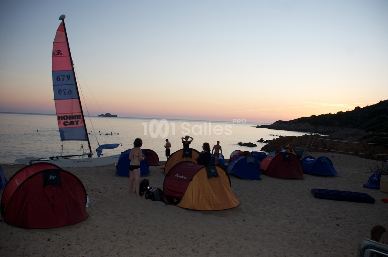 Des tentes installées sur une plage au coucher du soleil, avec des personnes près d'un voilier.