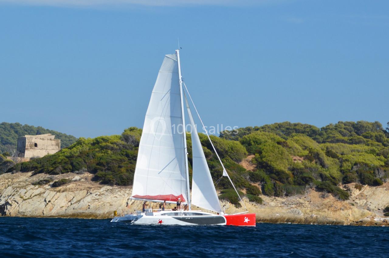 Catamaran à voile blanche naviguant près d'une côte rocheuse bordée de végétation et d'un bâtiment ancien.