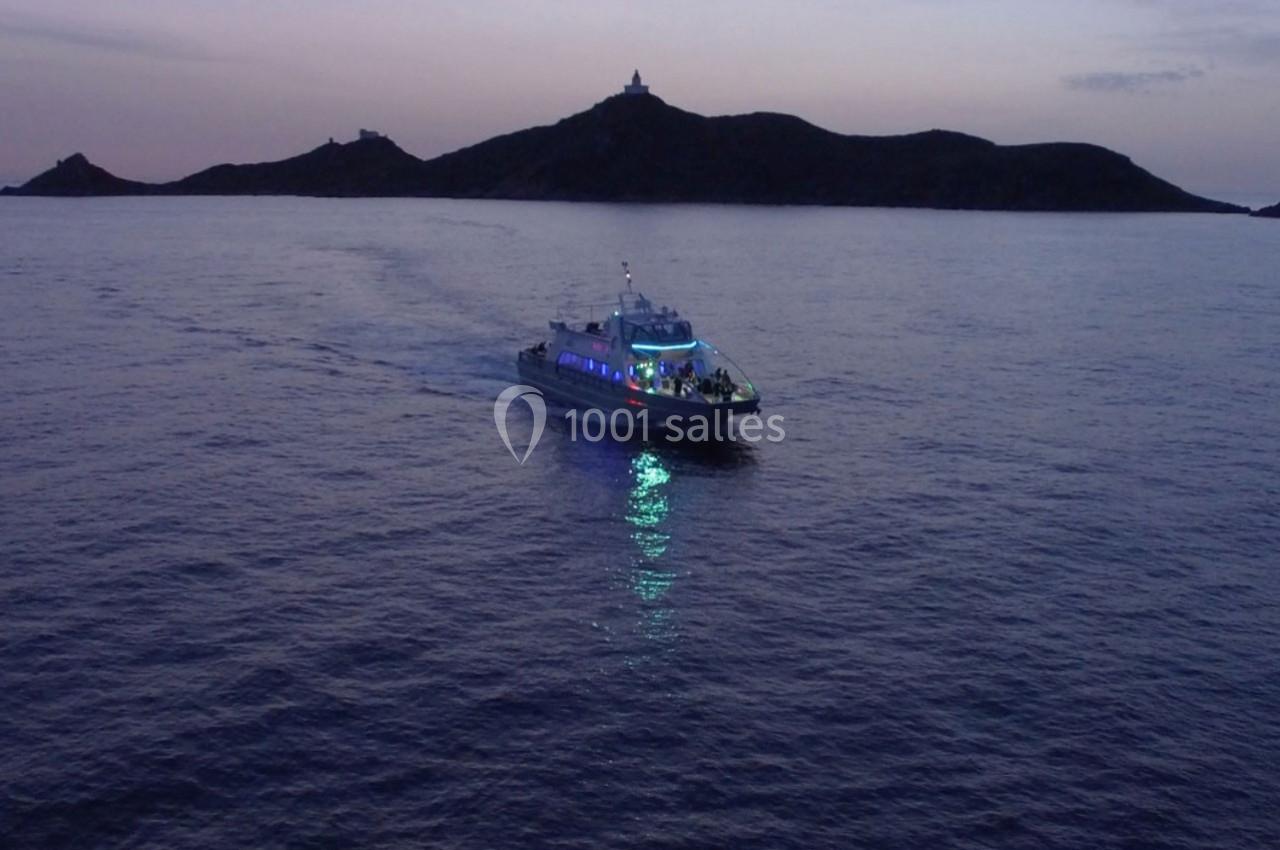 Un bateau éclairé navigue de nuit sur une mer calme, avec une île sombre en arrière-plan.