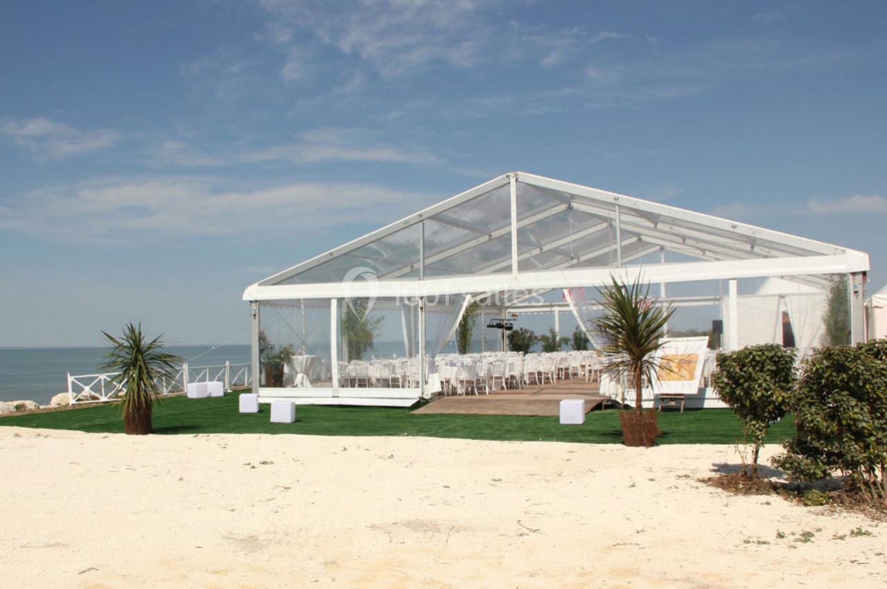 Tente transparente installée sur une plage, aménagée avec des chaises blanches et des plantes décoratives.