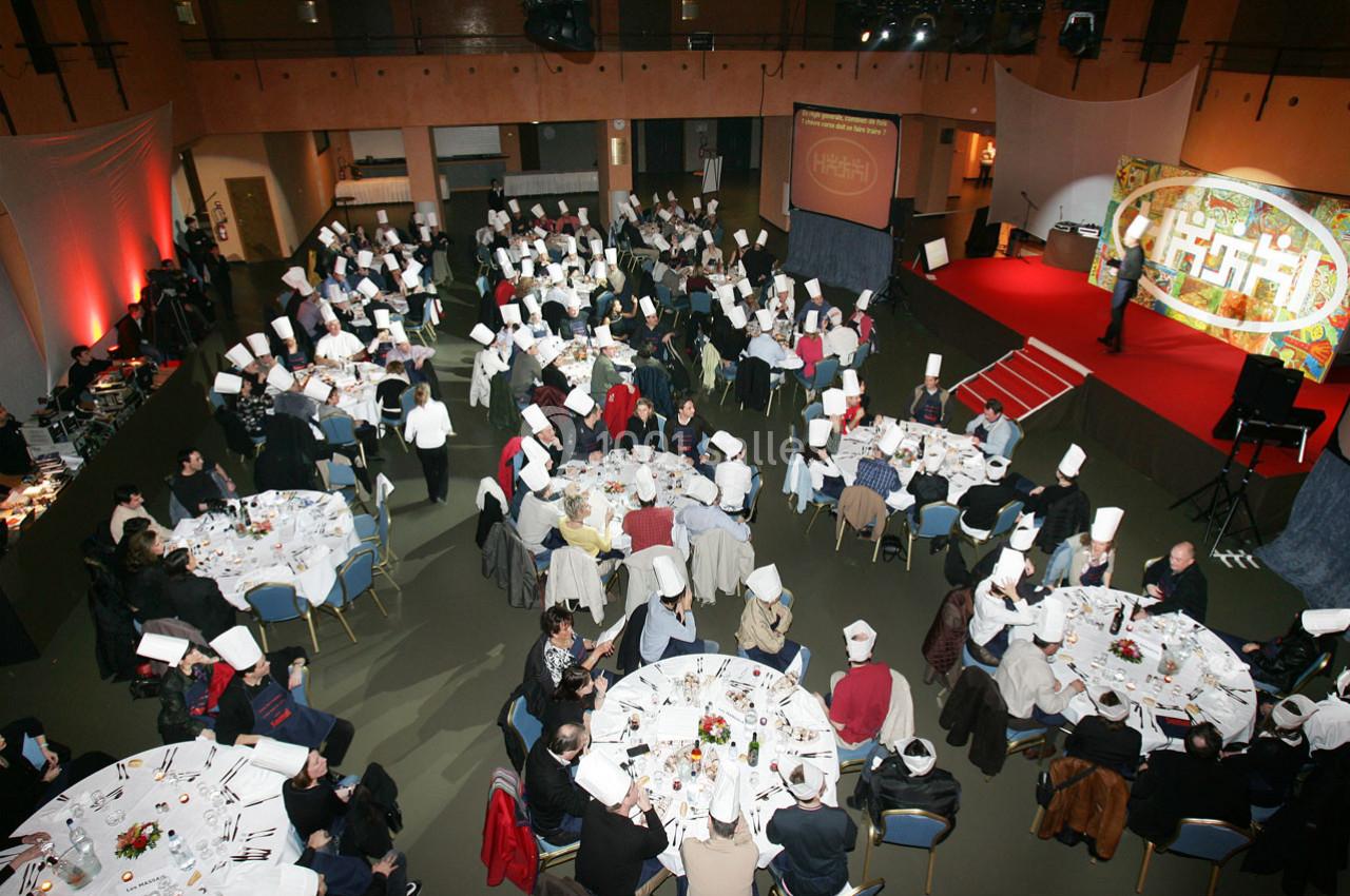 Salle de réception avec des tables rondes, des convives portant des toques blanches et une scène en arrière-plan.