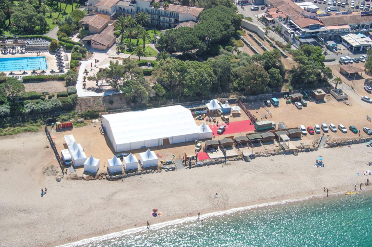 Vue aérienne d'une plage avec une grande structure blanche, des tentes, des voitures et des bâtiments en arrière-plan.