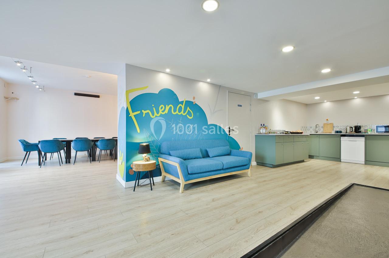 Espace lumineux avec canapé bleu, mur décoré ’Friends’, coin cuisine et table à manger en arrière-plan.