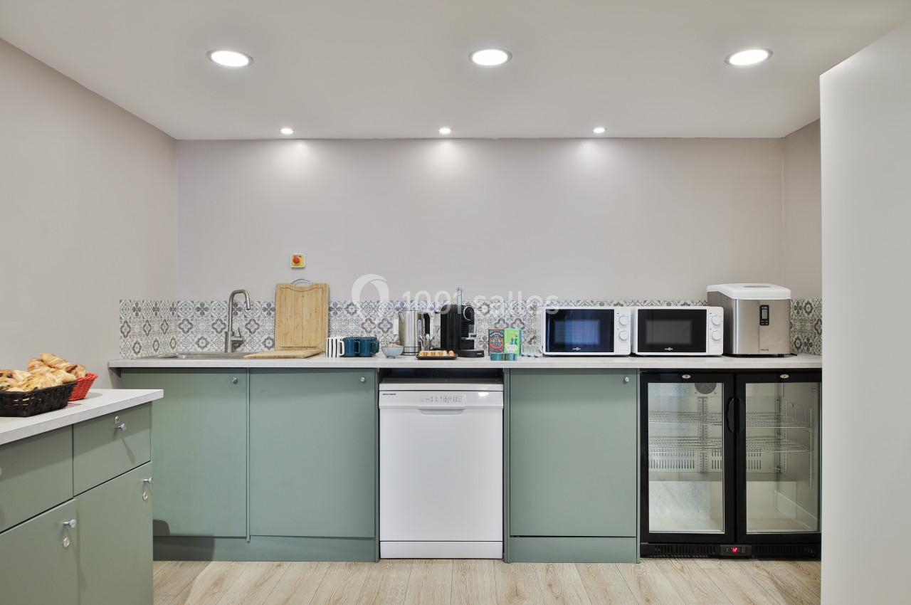 Cuisine moderne avec plan de travail, micro-ondes, lave-vaisselle, réfrigérateur, cafetière et panier de viennoiseries.