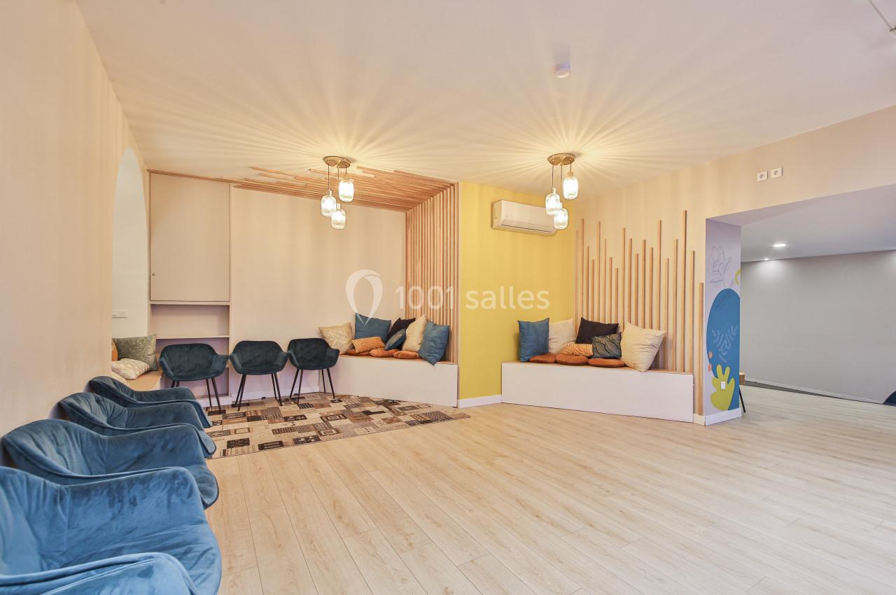 Salle lumineuse avec parquet clair, banquettes garnies de coussins colorés et chaises bleues disposées en rangée.