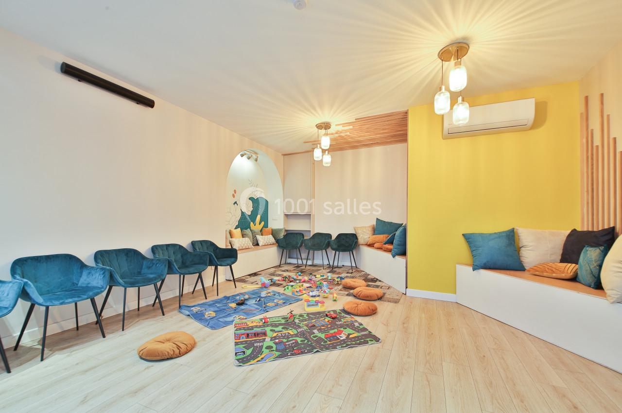 Salle lumineuse avec des sièges bleus, coussins colorés, tapis de jeu pour enfants et mur jaune décoratif.