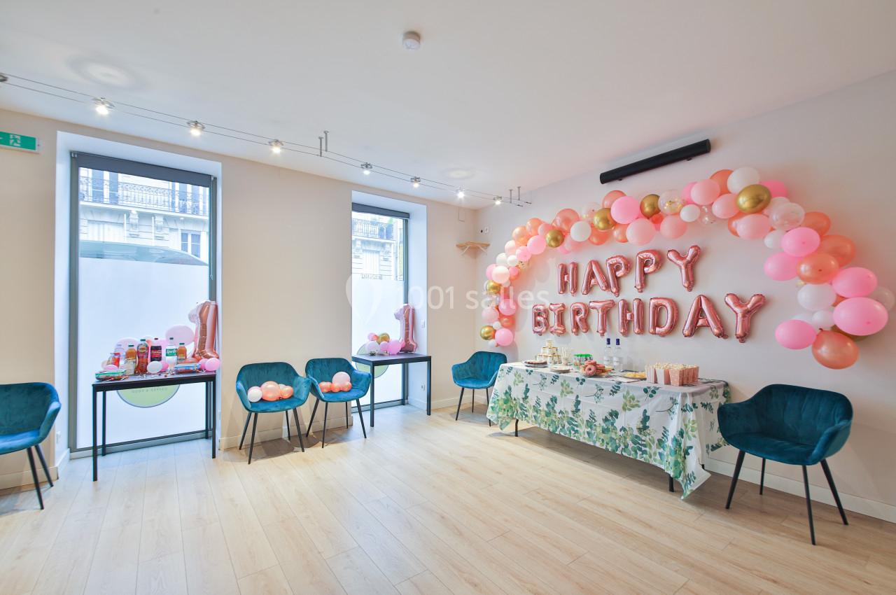 Salle décorée pour un anniversaire avec ballons roses et dorés, table garnie et chaises bleues.
