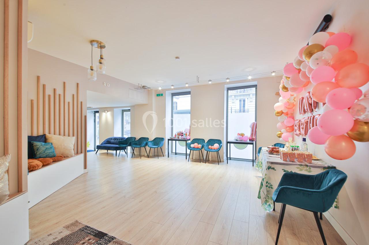 Salle lumineuse avec parquet, décorée de ballons colorés, tables dressées et fauteuils bleus près des fenêtres.