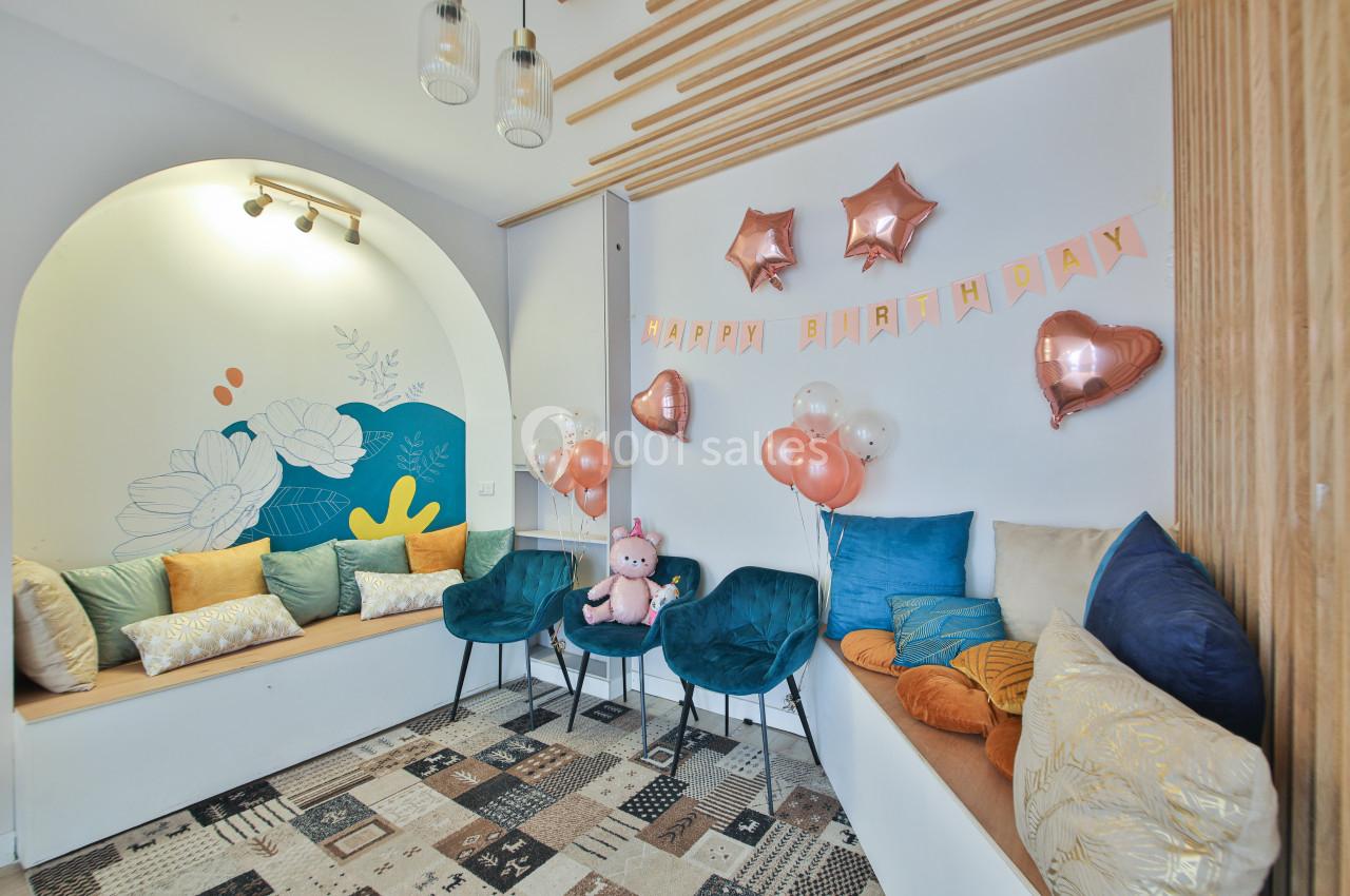 Salle décorée pour un anniversaire avec ballons roses, guirlande ’Happy Birthday’ et coussins colorés sur des banquettes.