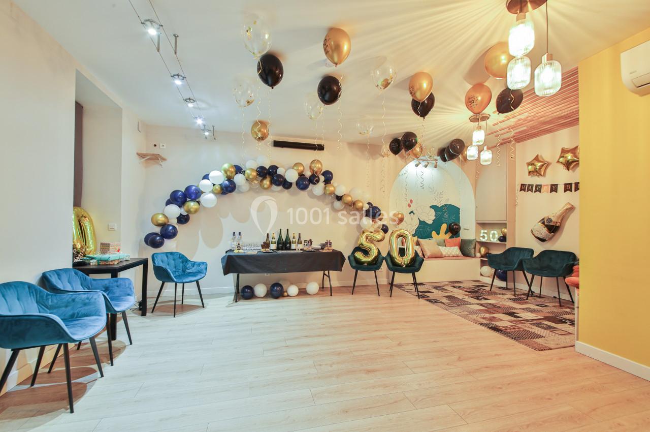 Salle décorée pour une fête avec ballons dorés, noirs et blancs, table avec boissons et fauteuils bleus.