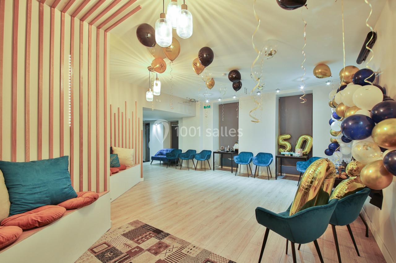 Salle décorée pour une fête avec ballons dorés, noirs et blancs, chaises bleues et coussins colorés.