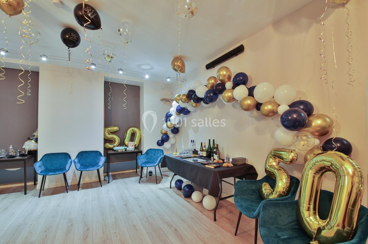 Salle décorée pour un 50e anniversaire avec ballons dorés, bleus et blancs, et table garnie de boissons et desserts.