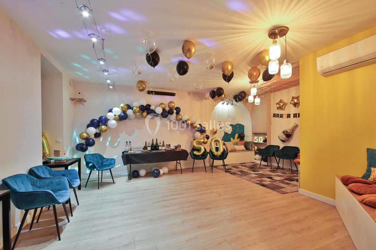 Salle décorée pour une fête avec ballons dorés, noirs et blancs, chiffre ’50’ en ballon et table avec boissons.
