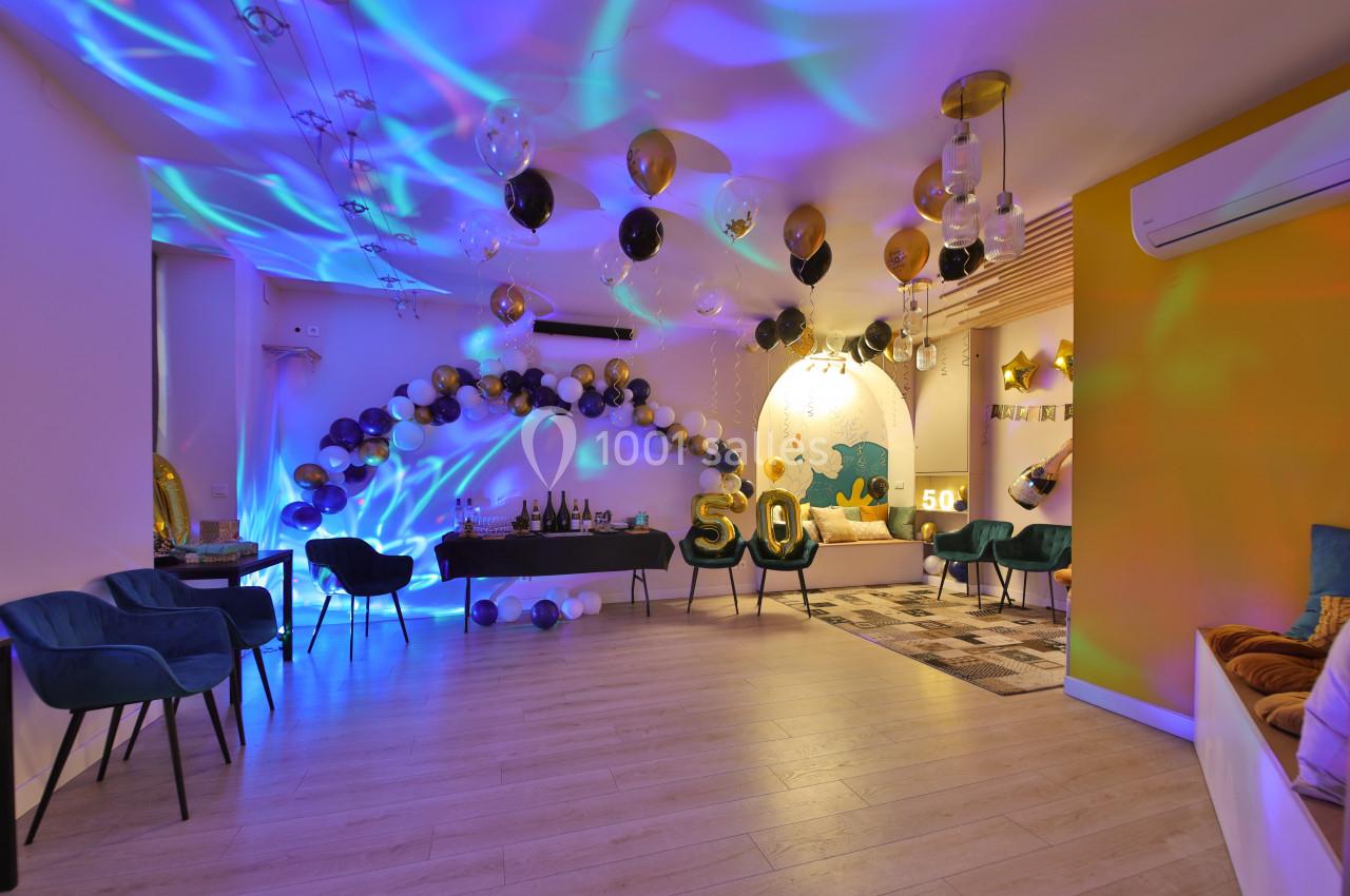 Salle décorée pour une fête avec ballons noirs et dorés, chiffre 50 en évidence, lumière colorée et mobilier moderne.