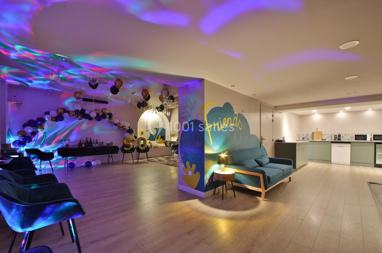 Salle décorée pour une fête avec ballons, éclairages colorés, coin salon et cuisine ouverte en arrière-plan.