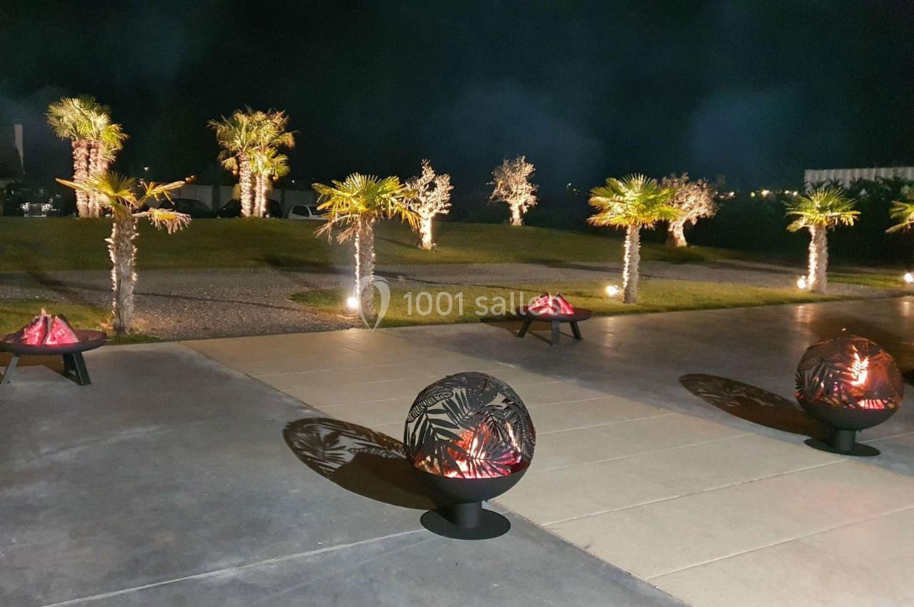 Espace extérieur nocturne avec palmiers éclairés et braseros décoratifs projetant des ombres sur le sol.