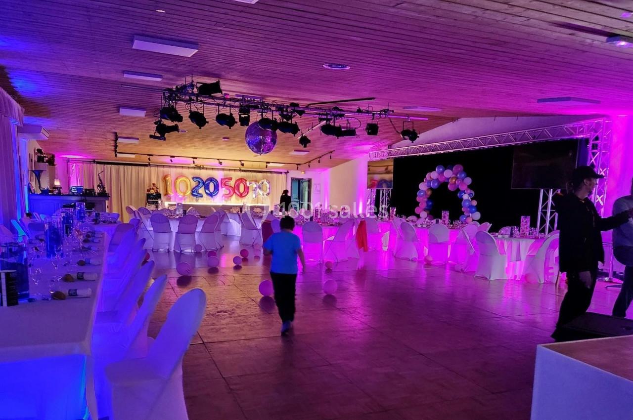 Salle de réception décorée avec des tables dressées, des ballons et un éclairage coloré, avec quelques personnes présentes.