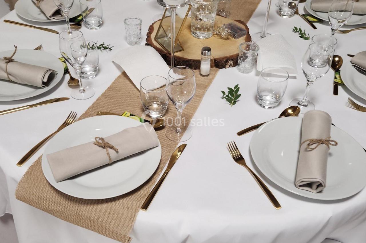 Table dressée avec une nappe blanche, couverts dorés, serviettes beige nouées, chemins de table en toile de jute et…
