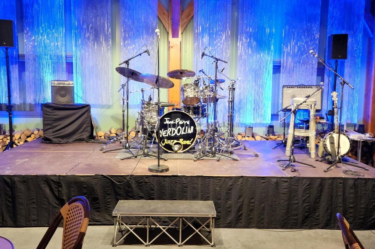 Scène de concert vide avec batterie, percussions et amplificateur, éclairée par des lumières bleues.