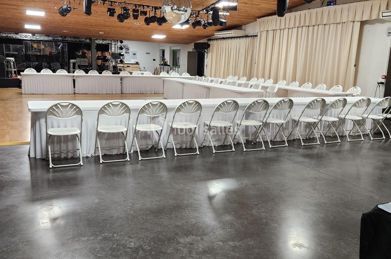Salle avec un sol en béton, tables disposées en U avec nappes blanches, chaises pliantes et éclairage suspendu.
