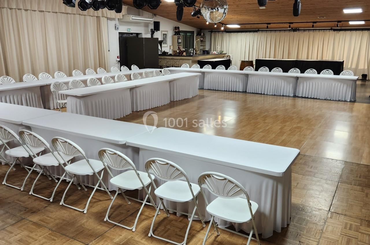 Salle de réception avec parquet, tables blanches disposées en U, chaises pliantes et éclairage suspendu.